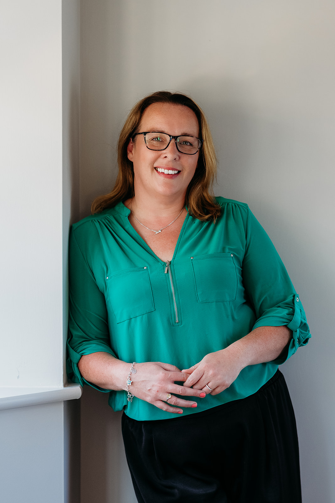 A smiling woman wearing glasses and a green blouse leans against a wall.