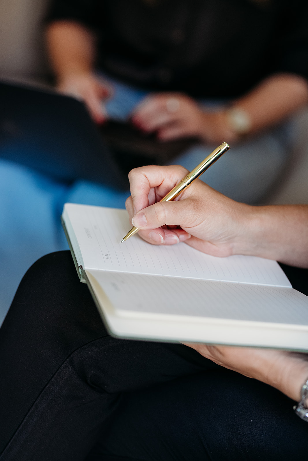 A person holds a gold pen and writes in an open notebook, with another person working on a laptop in the blurred background.