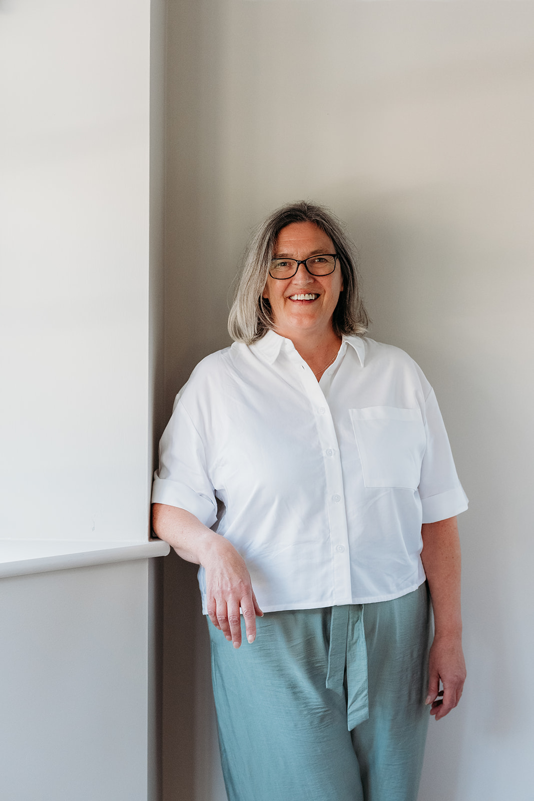 A smiling woman with shoulder-length grey hair and glasses wearing a white short-sleeved button-down shirt and light green trousers, leaning against a wall.